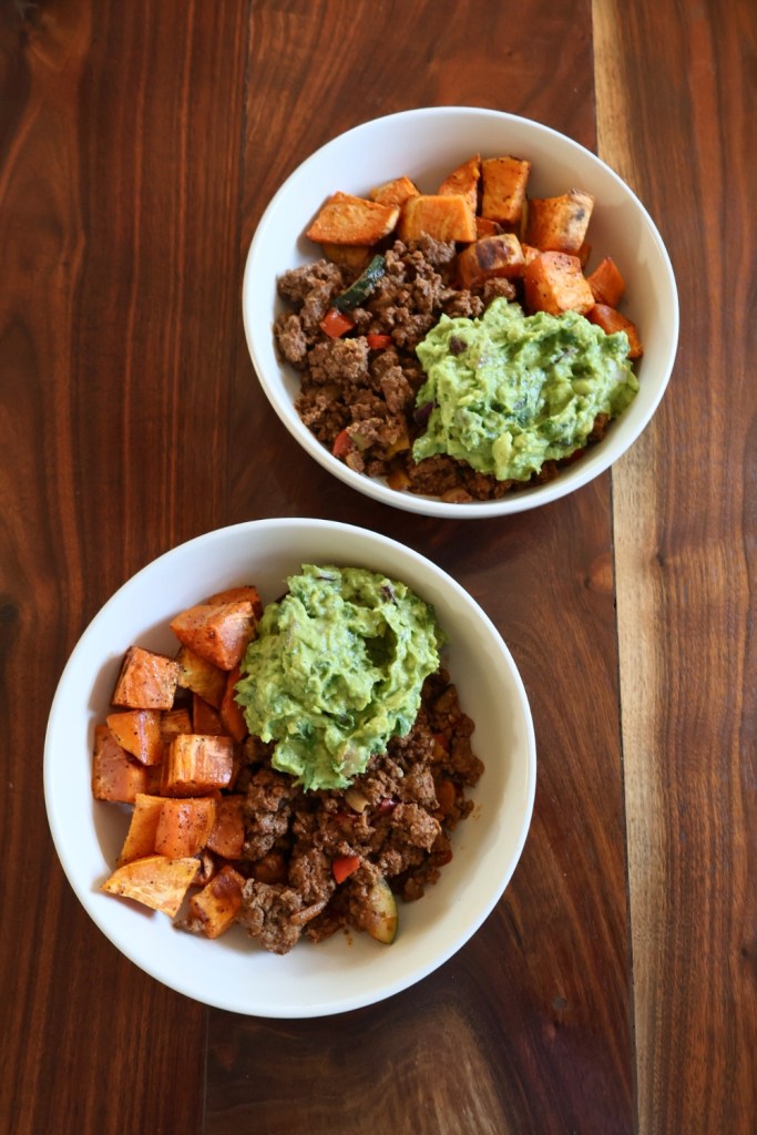 Colorful beef and sweet potato taco bowl topped with fresh guacamole, cilantro, and lime wedges, served in a white bowl.