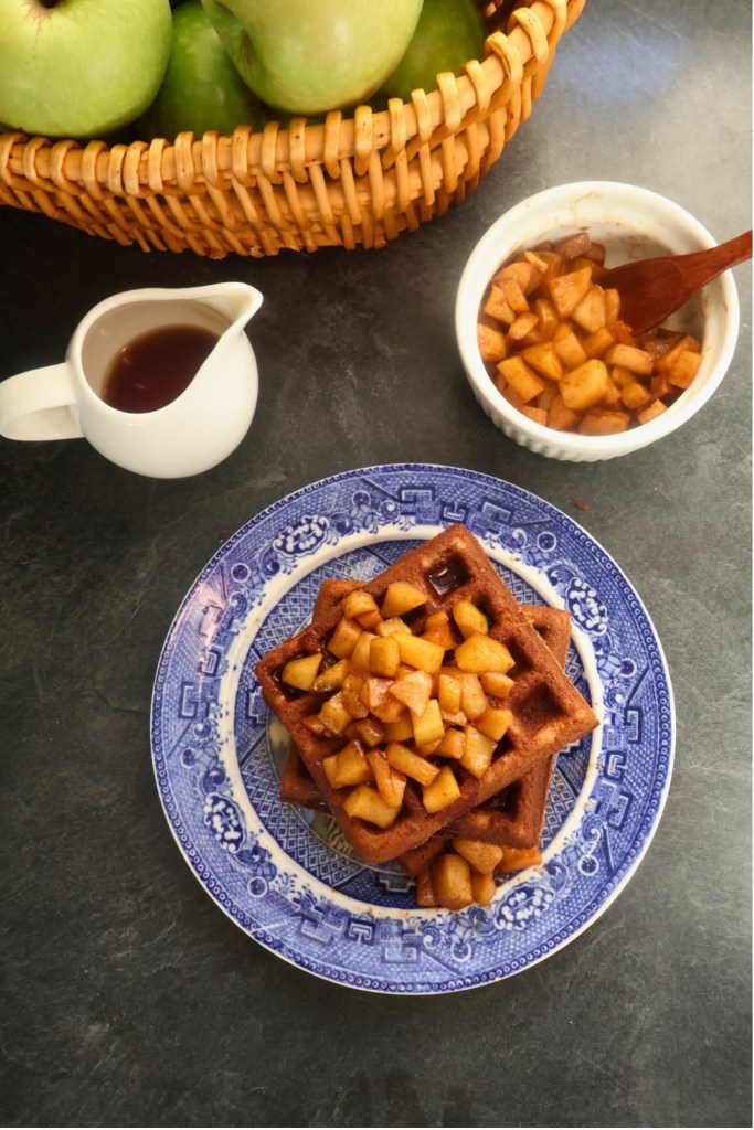 Overhead view of apple spice waffles served with cinnamon apples, maple syrup, and coffee on a rustic table.